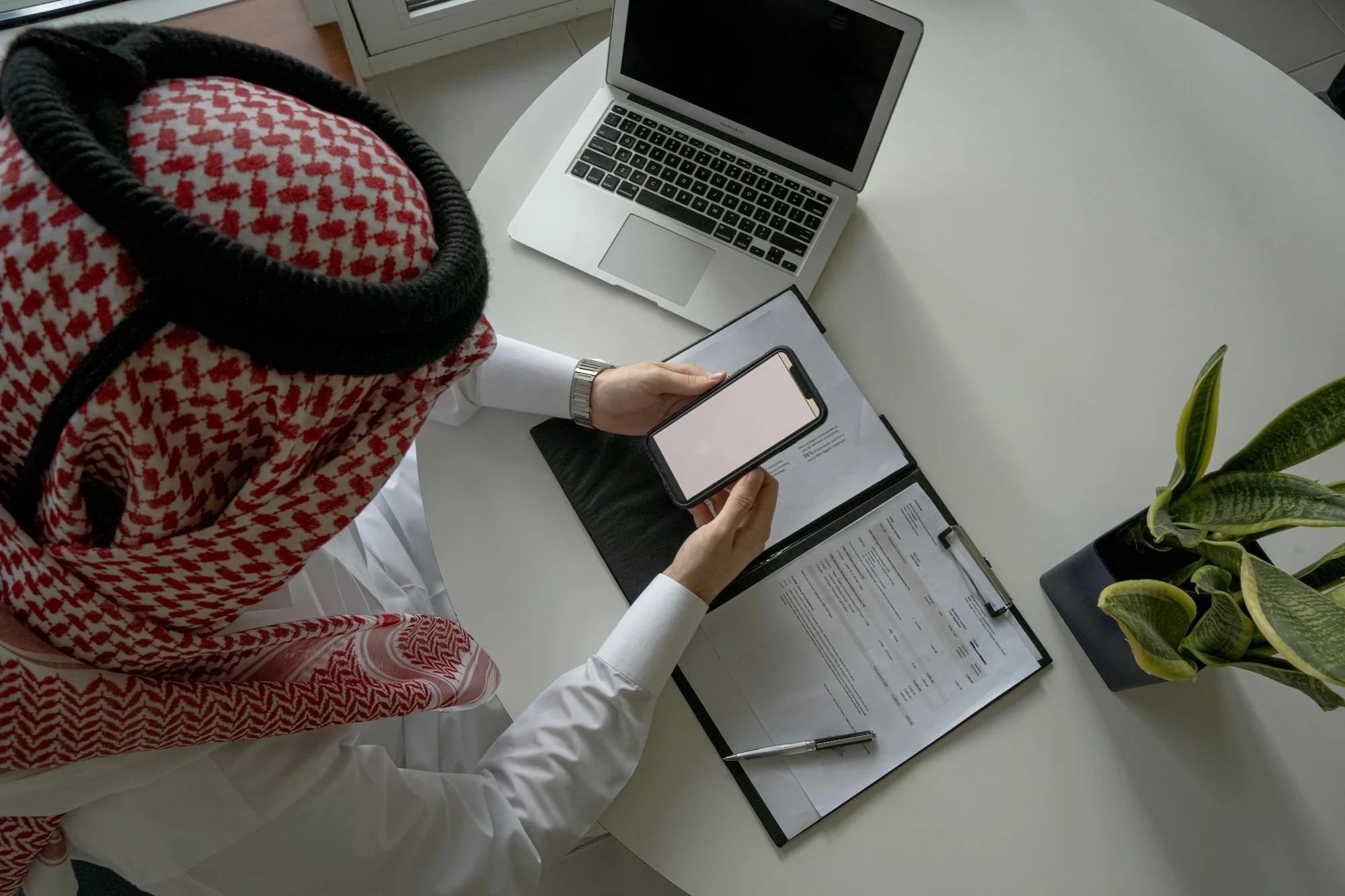 Saudi professional in a shemagh reviewing documents and a phone at a desk
